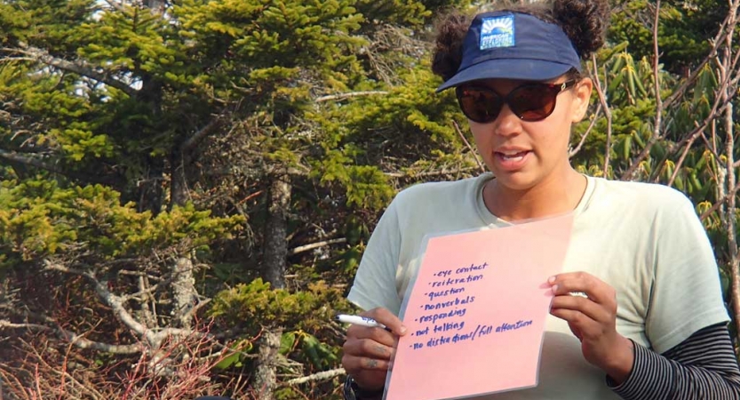 A person standing in a wooded area holds a pink piece of paper with writing. 